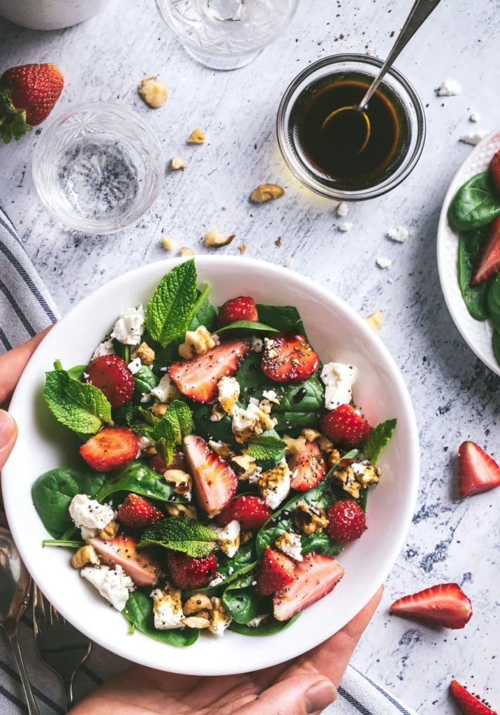 A white bowl filled with fresh sliced strawberries, mint leaves, spinach, shredded cheese, and walnuts, creating a colorful and nutritious salad.