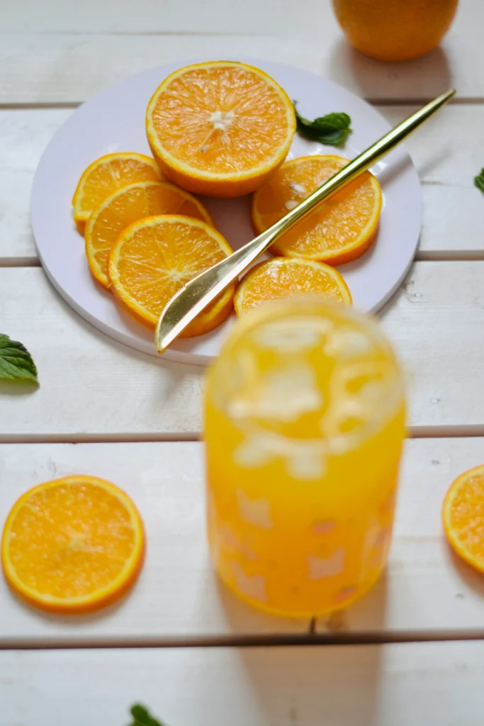 "A clear glass of fresh orange juice on a sunlit kitchen counter, surrounded by orange slices and a water pitcher, symbolizing hydration and health.