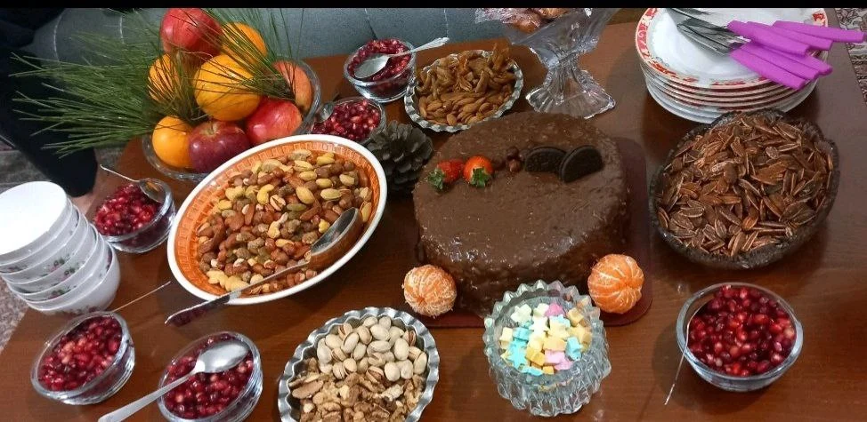 Modern Yalda Night table with pistachios, cake, mixed nuts, baslogh sweets, apples, oranges, and individual bowls of pomegranate seeds on a wooden table
