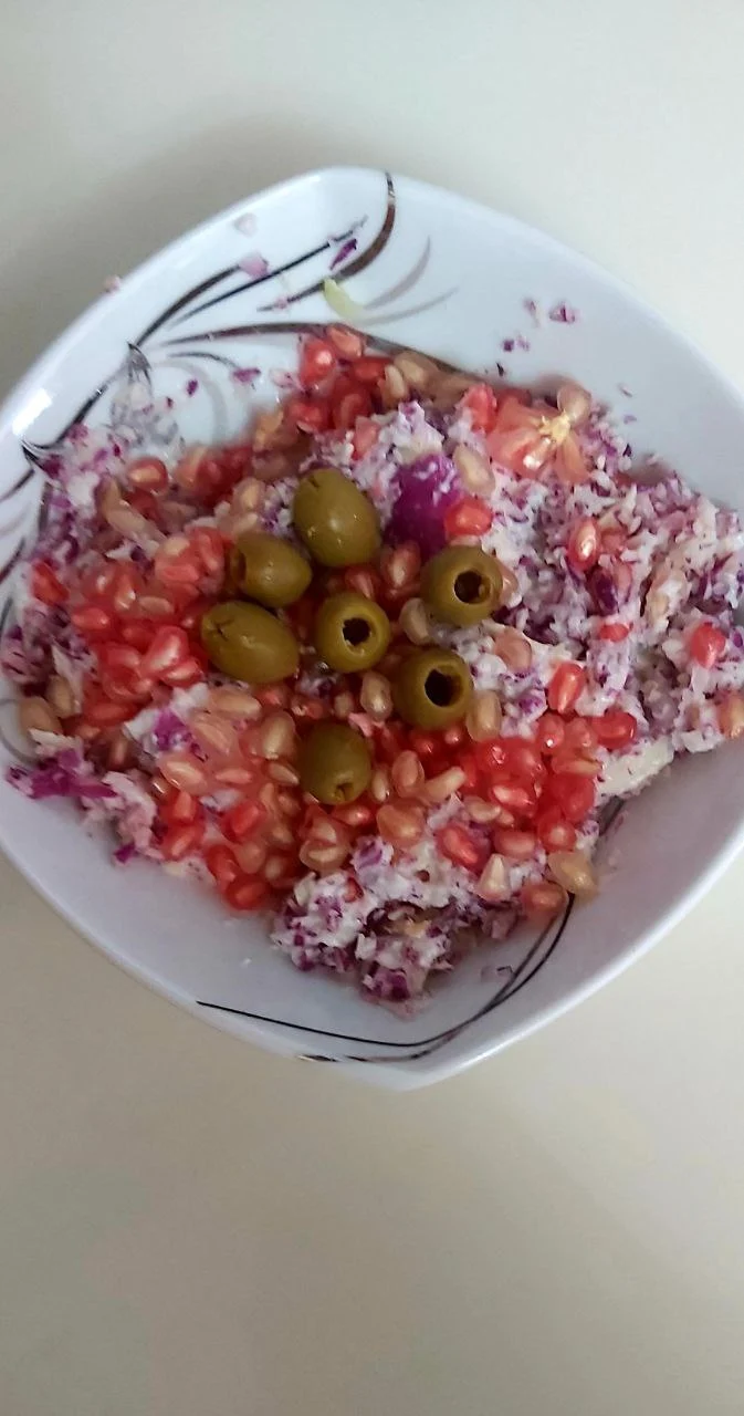 A white porcelain bowl filled with shredded white and red cabbage, garnished with pomegranate seeds and pitted olives, placed on a light surface.