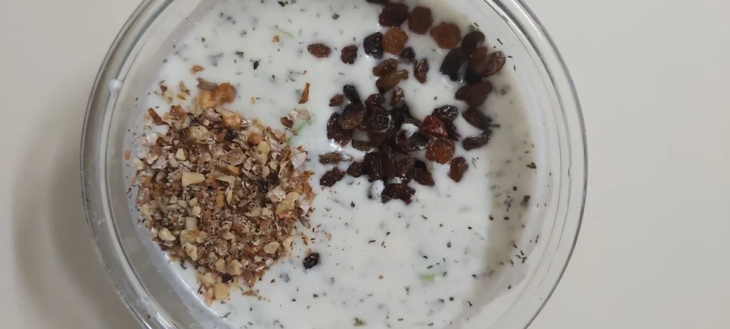 A crystal bowl filled with a yogurt and cucumber mix, garnished with dried mint, powdered rose, chopped walnuts, and raisins.