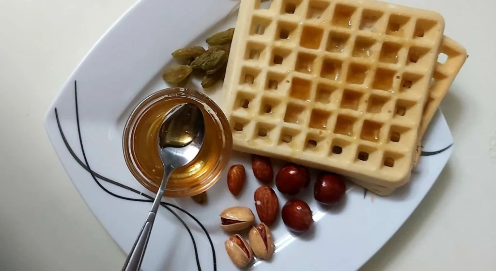 A square china plate with diet waffles, pistachios, hazelnuts, raisins and almonds A small container of honey with a spoon.