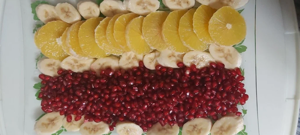 A crystal dish featuring a row of vibrant orange slices, a row of red pomegranate seeds, and banana slices placed between the rows and along the sides. These colorful fruits represent healthy snack options for weight loss.