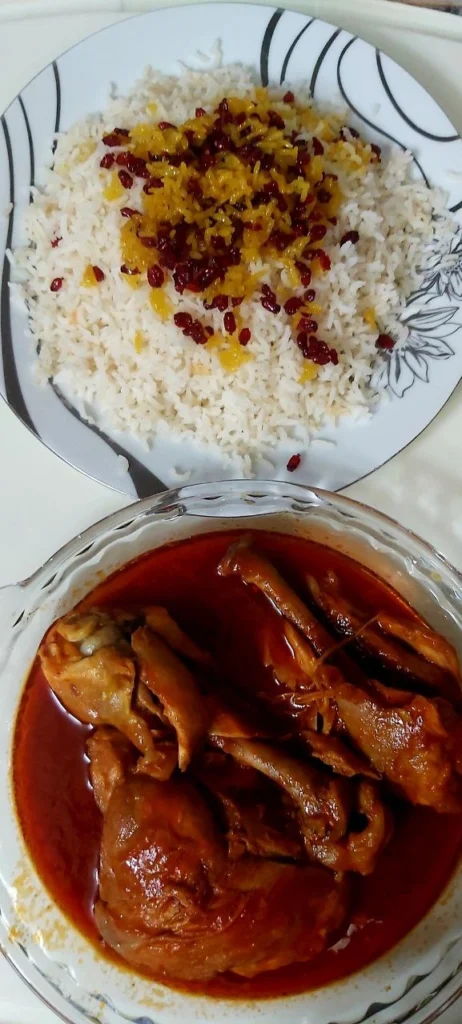 A plate of Persian rice topped with saffron and barberries, served with pieces of chicken in tomato sauce.