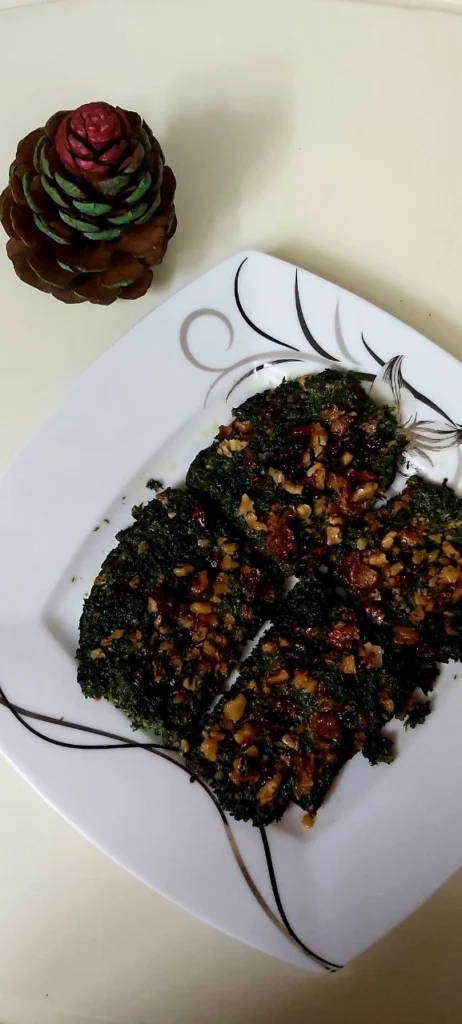 A square Chinese plate with a vegetable coco appetizer topped with walnuts and barberries is next to a green and brown pine cone.
