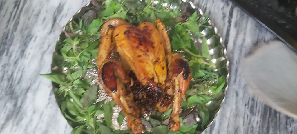 A whole roasted chicken in a round steel tray, surrounded by roasted vegetables like carrots, potatoes, and broccoli, set on a wooden table.