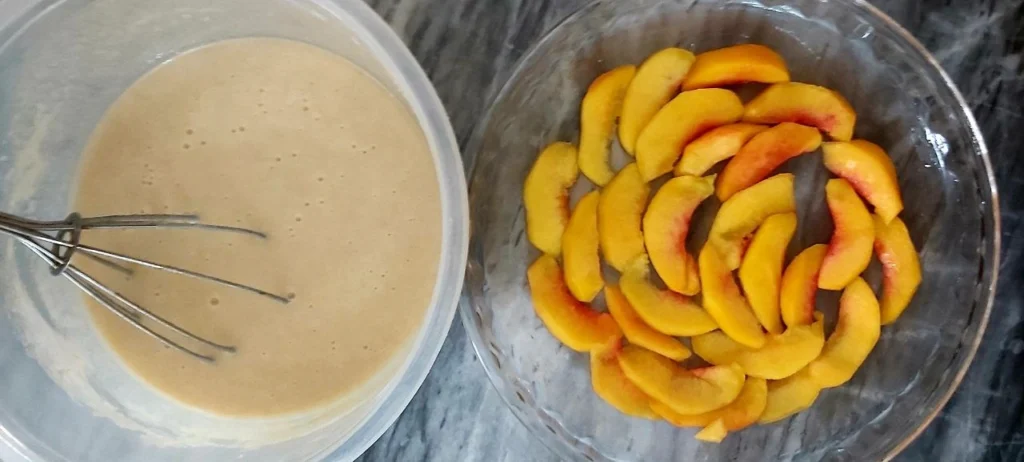 Cake batter in mixing bowl with electric mixer next to Pyrex dish lined with peach slices