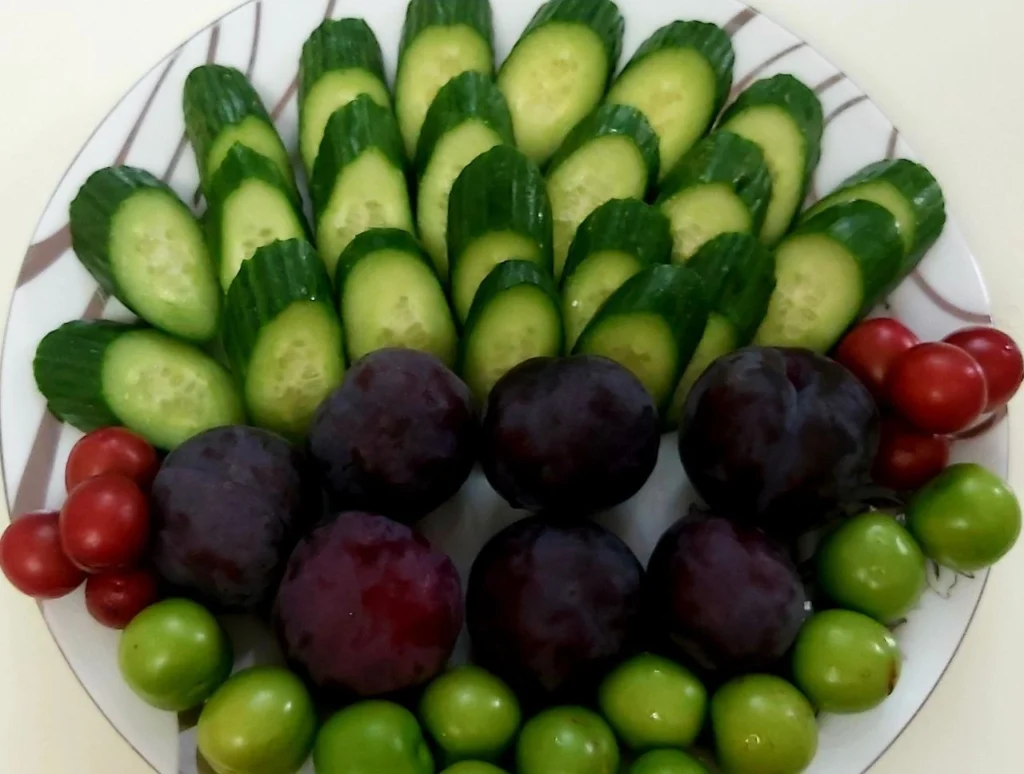 Cucumbers and green and red plums arranged in a white porcelain dish, showcasing their freshness and vibrant colors