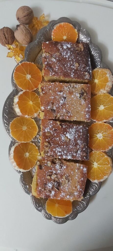 Four slices of golden orange cake arranged neatly on a shiny nickel silver tray, surrounded by fresh round orange slices, with a few walnuts scattered outside the tray on a rustic surface.