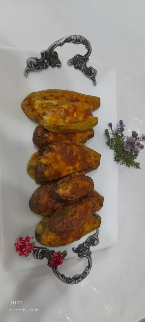 Fried, peeled eggplant pieces stuffed with minced meat and onions, topped with melted pizza cheese. The dish is arranged on a white porcelain plate with a silver metal handle, placed on a white countertop. A small bunch of chamomile flowers rests beside the plate.