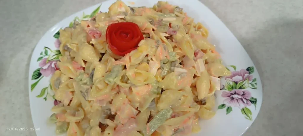 A plate of colorful pasta salad featuring various shapes in yellow and green, garnished with a decorative red flower made of tomatoes.