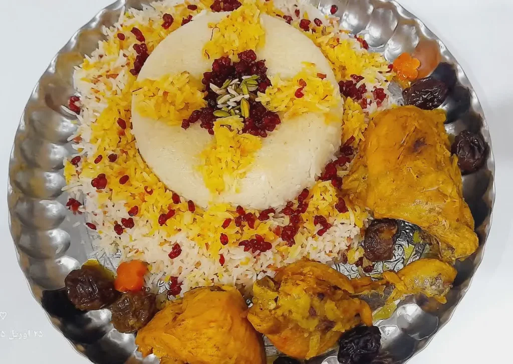 A beautifully molded round saffron rice dish topped with barberries, surrounded by golden fried chicken pieces, stewed prunes, and carrots, served on a rustic wooden background – a traditional Persian presentation