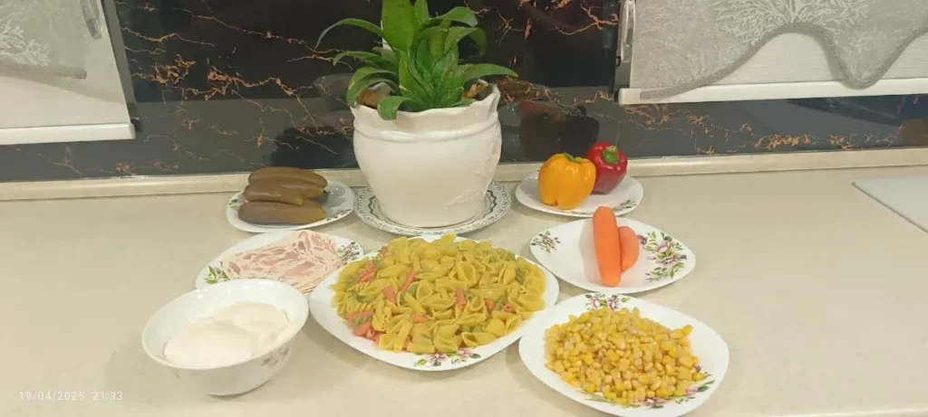 A visually appealing arrangement of pasta salad ingredients, featuring six small plates of carrots, red and yellow bell peppers, pickled cucumbers, mayonnaise, cooked corn, and sausage slices, surrounding a large plate of colorful pasta in yellow and red sauce. A vase of natural flowers sits in the background.