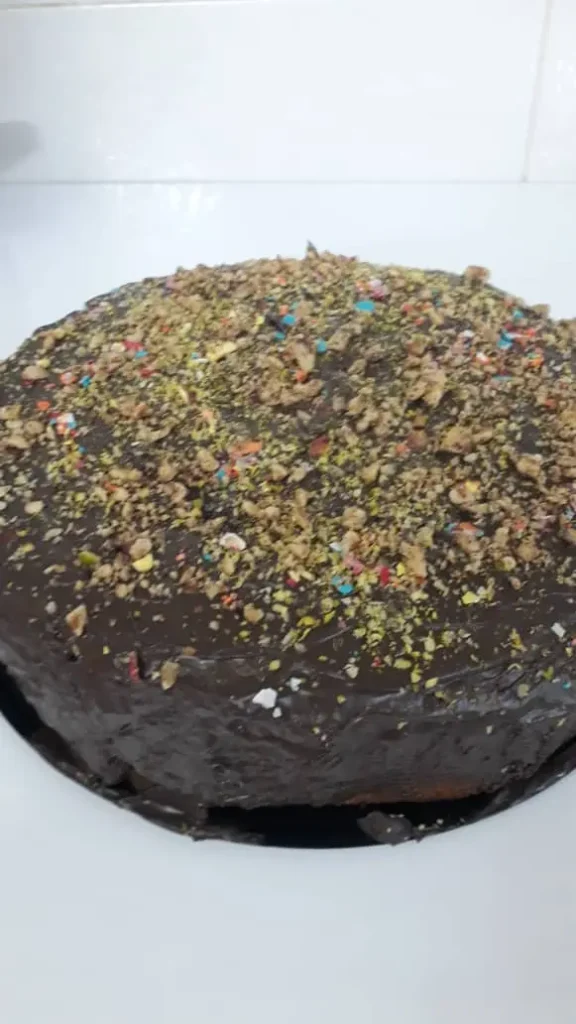 A beautifully crafted 25 cm round chocolate cake, adorned with vibrant green pistachio and rich walnut powder, artistically sprinkled with colorful food coloring.