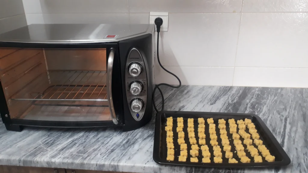 A pea-shaped cookie with a delicate 4-petal flower design rests on a black tray next to a toaster, which is illuminated from the inside.