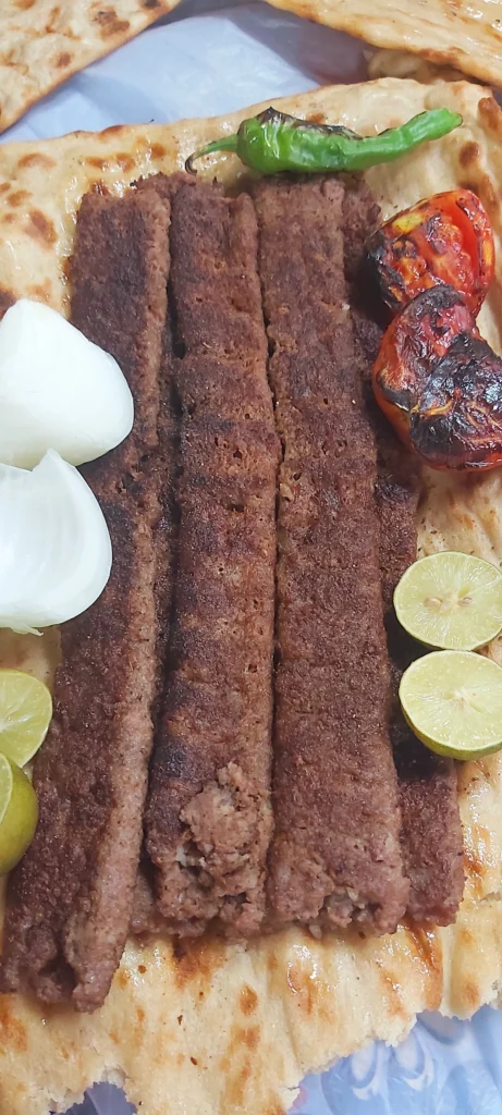 Grilled Persian Koobideh kebabs served on traditional Sangak bread, topped with charred tomatoes, onion slices, lemon wedges, and fresh herbs.
