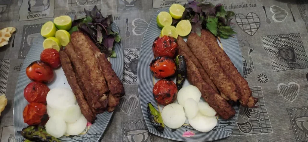 Koobideh kebab on platters with lemon wedges, roasted tomatoes, grilled peppers, and fresh basil.