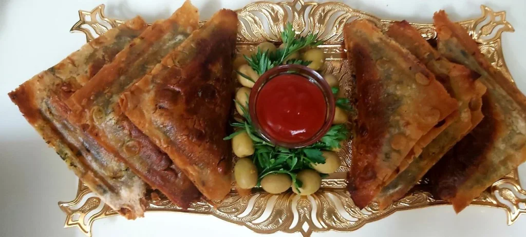Golden fried samosas placed in a metallic golden tray, with a small bowl of ketchup surrounded by parsley and olives.