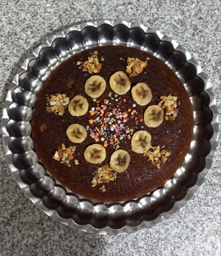 A round chocolate cake in a round steel dish, decorated with banana slices, chopped walnuts, and colorful sprinkles in the center.