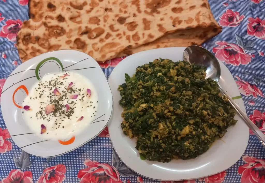 A plate of green spinach borani next to a white bowl of yogurt garnished with pink rose petals and green mint leaves