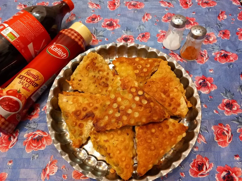 Triangular sambusas arranged on a large stainless steel tray, served with salt, pepper, soda, and various dipping sauces.