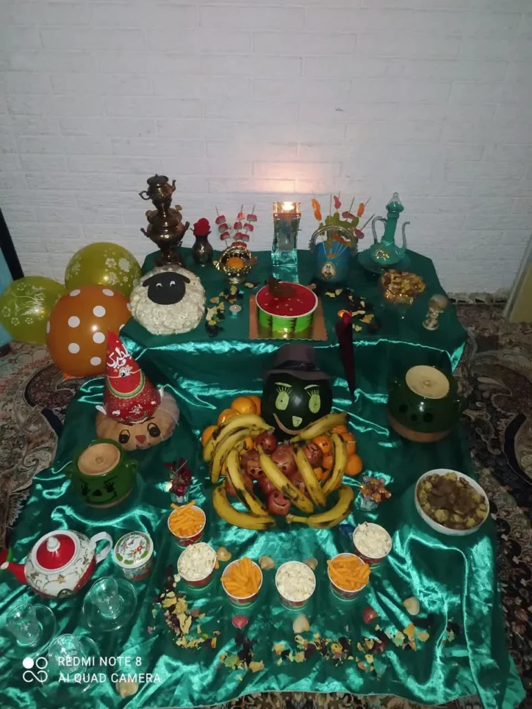 A festive Shab-e Yalda spread arranged on a green tablecloth. The spread includes a pumpkin, popcorn, nuts, a cake, and fruits such as bananas, pomegranates, oranges, and various snacks.