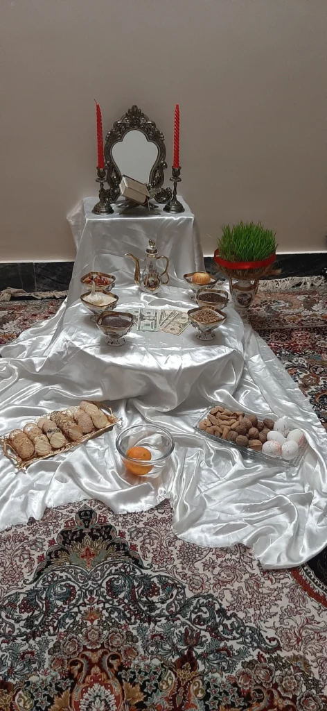 A traditional Haft-Sin table with seven symbolic items arranged on white satin. The top tier features a Quran, a mirror, and two red candles. The second tier displays the seven ‘S’ items: sumac, samanu, senjed,see, serkeh, sib, and sekkeh, representing ancient Persian heritage. Below, elements of water, fruits, sweets, and eggs symbolize purity, abundance, and new life.”